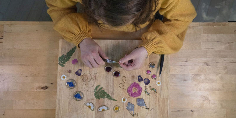 Person working on a wooden board with various small jewelry components, dried flowers, and designs.